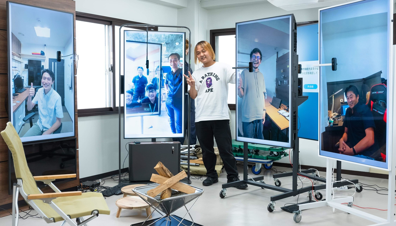 A man in a white T‑shirt stands between four large video screens showing remotely connected teammates in a tech workspace, making a peace sign.
