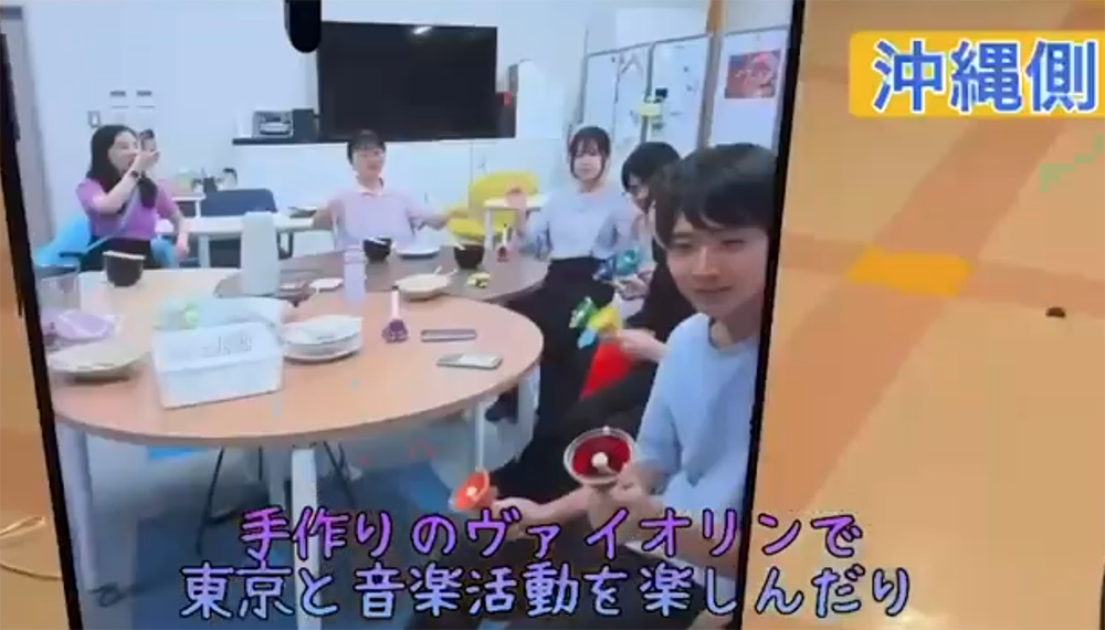 Six people sit around a round table in a bright room, with snacks and drinks, while a large screen looms in the background; Japanese subtitles appear at the bottom of the frame.
