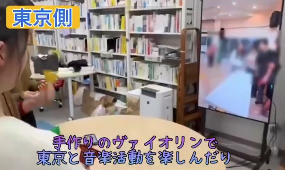 Person sits at a round table in a bookshelf-filled room; a large monitor on the right displays a blurred group video call.